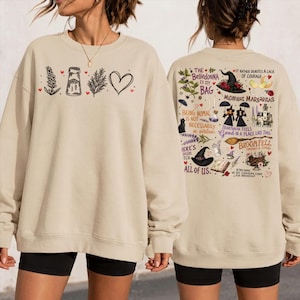 May include: A beige crewneck sweatshirt featuring a front graphic of a lavender sprig, salt shaker, rosemary sprig, and heart. The back displays various witch-themed illustrations and text including Midnight Margaritas and Broom Fell.