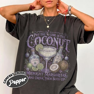 May include: A charcoal gray short-sleeve t-shirt featuring a graphic print with the text Put the lime in the coconut, Midnight Margaritas, and drink them both up. The design includes illustrations of a margarita glass, tequila bottles, and coconuts.