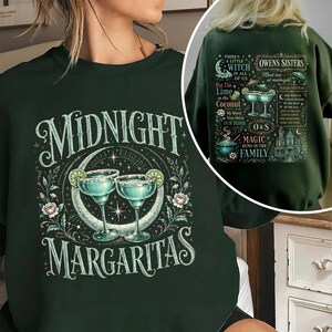 May include: A dark green crewneck sweatshirt featuring a front graphic of two margarita glasses with lime slices, a crescent moon, and the text "MIDNIGHT MARGARITAS." The back graphic displays "OWENS SISTERS" with various witch-themed illustrations and text.