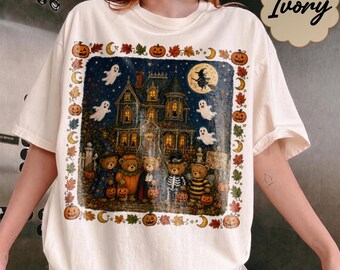 May include: An ivory short-sleeve t-shirt featuring a rectangular graphic of a haunted house at night with a full moon, flying ghosts, and five teddy bears in Halloween costumes. The border includes pumpkins, autumn leaves, and crescent moons.