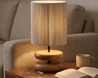 May include: A table lamp with a cylindrical shade wrapped in light gray vertical threads, featuring a base composed of two stacked, smooth, rounded wooden discs connected by a thin black metal rod, with the text VELLARO LAMP visible.