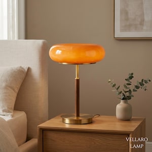 May include: A table lamp featuring a smooth, translucent orange glass shade shaped like a flattened sphere. The lamp has a slender central stem made of dark brown wood, supported by a circular brushed brass base and matching brass accents.