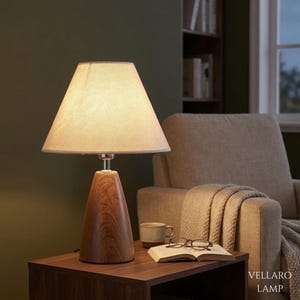 Op de afbeelding: Een tafellamp met een conische houten voet met een zichtbare houtnerf en een lichtgekleurde, taps toelopende stoffen lampenkap. De lamp geeft een warm licht dat de omgeving verlicht. De tekst VELLARO LAMP is zichtbaar in de hoek.