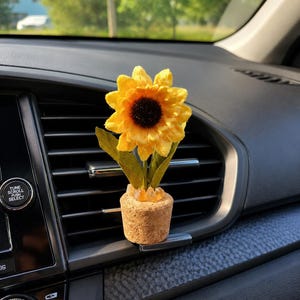 May include: A sunflower car air freshener featuring a bright yellow flower with a dark brown center, green leaves, and a small cork base filled with orange crystal chips, attached to a black plastic vehicle air vent clip.