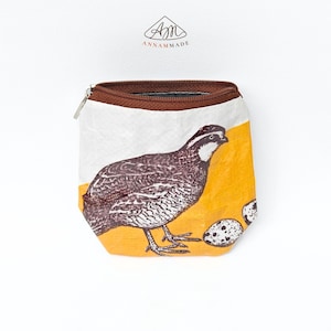 May include: A small pouch with a brown zipper closure featuring a printed graphic of a quail and two speckled eggs against a white and yellow background. The logo ANNAMMADE is printed above the pouch in black and red text on a white surface.