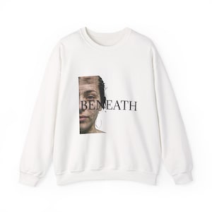 Puede incluir: Una sudadera blanca de manga larga y cuello redondo con un gráfico rectangular en la parte delantera. La imagen muestra la mitad izquierda de un rostro parcialmente sumergido en agua, con la palabra BENEATH impresa en letras mayúsculas negras.