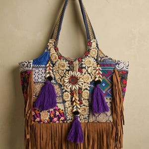 Op de afbeelding: Een tote bag met een patchworkontwerp van blauwe, roze en gouden stoffen met ingewikkeld borduurwerk. De tas is versierd met kaurischelpen, een centrale spiegel en drie paarse kwastjes. Een lange bruine franje hangt aan de onderkant.