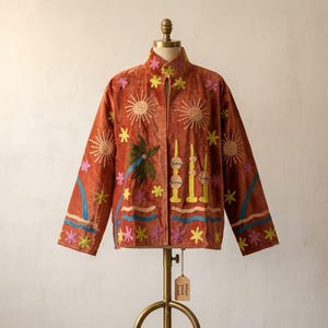 以下が含まれることがあります： ハイカラーと長袖が特徴の錆色のベルベットジャケット。太陽、ヤシの木、キャンドル、花の刺繍が黄色、ピンク、緑、青の色合いで施されており、マネキンにディスプレイされています。