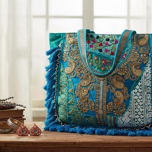 Può includere: Una borsa tote color verde acqua con intricati ricami dorati, motivi floreali e nappe blu decorative lungo i lati e il fondo. La borsa presenta due manici in tessuto coordinato e una forma rettangolare con pannelli in tessuto testurizzato.