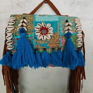 Puede incluir: Un bolso rectangular con un mosaico de tela azul verdoso y dorada con bordados intrincados. El bolso está decorado con conchas de cauri blancas, grandes borlas de hilo azul y largas tiras de flecos de piel sintética marrón en los lados.