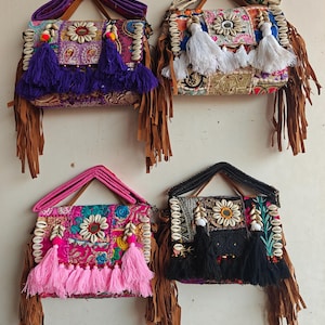 May include: Four rectangular shoulder bags featuring patchwork fabric, cowrie shell accents, and long brown fringe. Each bag has a top handle, a flap closure, and large decorative tassels in purple, white, pink, or black hanging from the front.