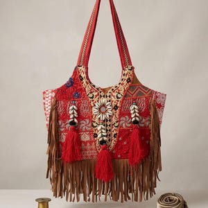 Könnte beinhalten: Eine rote Tragetasche mit besticktem Patchwork, braunen Fransen aus Wildlederimitat und Details aus natürlichen Kaurimuscheln. Die Tasche hat zwei rote Schulterriemen und ist mit roten Quasten verziert. Der Text unten lautet: Artisanal Red Tote with Embroidered Patchwork, Faux-suede Fringe, and Natural Cowrie Shell Detail.