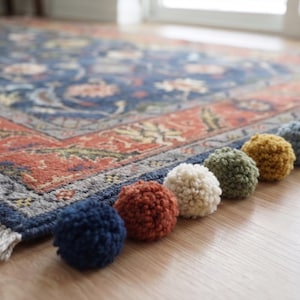 May include: A rectangular area rug featuring a dark blue center with intricate floral patterns in orange, cream, and green, bordered by a wide orange frame. A row of multi-colored yarn pom-poms in navy, rust, cream, olive, mustard, and grey lines the edge.