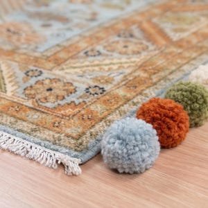 May include: A rectangular area rug featuring a light blue and tan floral pattern with a white fringe edge. A row of five fuzzy yarn pom-poms in light blue, burnt orange, olive green, cream, and dark charcoal is attached to the rug corner.