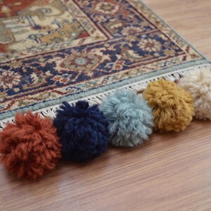 May include: A row of five round yarn pom-poms in rust, navy blue, dusty blue, mustard yellow, and cream colors. The pom-poms are arranged in a line on a wooden surface in front of a patterned area rug with red, blue, and beige geometric designs.