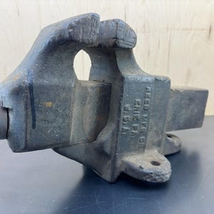 May include: A heavy-duty cast iron bench vise with a dark gray, weathered finish. The side of the metal body features embossed text reading REED MFG CO ERIE PA U.S.A. The tool includes a threaded handle mechanism and a wide, stationary base.
