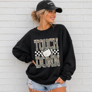 Könnte beinhalten: Ein schwarzes Langarm-Sweatshirt mit dem Text TOUCH DOWN in einem Tarnmuster und einer weißen Football-Grafik in der Mitte. Das Sweatshirt wird mit hellblauen Jeansshorts und einer schwarzen Baseballkappe mit dem Text Nashville kombiniert.