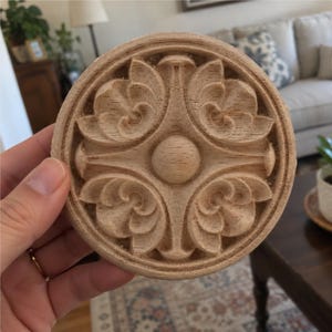May include: A circular light-colored wooden medallion featuring a carved relief design. The center displays a smooth sphere surrounded by four symmetrical floral-inspired motifs, all enclosed within a raised circular border frame.