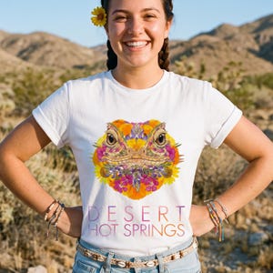 May include: A white short-sleeved t-shirt featuring a central graphic of a lizard head composed of colorful desert flowers in shades of yellow, orange, and purple. Below the floral lizard graphic, the text reads DESERT HOT SPRINGS.