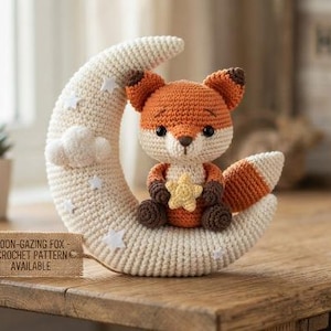 May include: A crocheted orange and cream fox plush toy sitting on a cream-colored crescent moon decorated with small white stars and a cloud. The fox holds a small yellow star. A rectangular tag reads MOON-GAZING FOX - CROCHET PATTERN AVAILABLE.