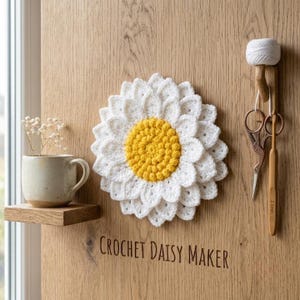 May include: A white and yellow crocheted daisy flower, a ceramic mug, a pair of metal scissors, and a wooden crochet hook. The text CROCHET DAISY MAKER is printed on the surface. The daisy has layered white petals and a textured yellow center.