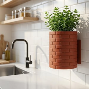 Puede incluir: Una maceta cilíndrica de pared con textura de ladrillo rojo, que contiene una frondosa planta verde. La maceta está fijada a una pared de azulejos blancos sobre una encimera de cocina, situada cerca de un grifo negro y un fregadero.