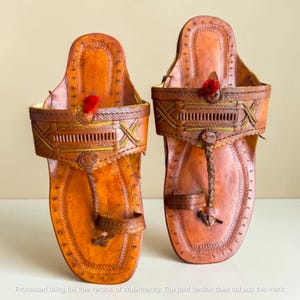 May include: A pair of tan leather sandals featuring intricate hand-stitched detailing, a braided toe post, and a decorative red pom-pom accent on the wide upper strap. The footwear includes yellow thread embroidery and a structured sole design.