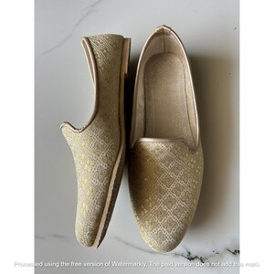 Pu&ograve; includere: Un paio di scarpe slip-on con una base in tessuto beige chiaro e un motivo floreale dorato ripetuto. Le scarpe presentano una punta arrotondata, una linea superiore bassa e un profilo sottile. La fodera interna &egrave; di colore beige chiaro.