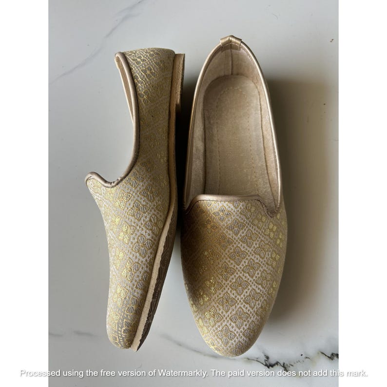 Pu&ograve; includere: Un paio di scarpe slip-on con una base in tessuto beige chiaro e un motivo floreale dorato ripetuto. Le scarpe presentano una punta arrotondata, una linea superiore bassa e un profilo sottile. La fodera interna &egrave; di colore beige chiaro.