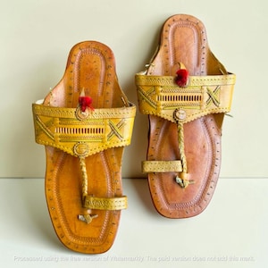 May include: A pair of tan leather sandals featuring a toe post, a wide strap across the instep with intricate stitched patterns, and small red decorative tassels. The sandals have a flat sole with detailed edge stitching and a square toe design.