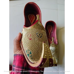 May include: A pair of gold-colored slip-on shoes featuring intricate metallic embroidery in paisley patterns with small red, blue, and green accents. The shoes have a pointed, upturned toe, a deep red interior lining, and a structured, flat sole design.