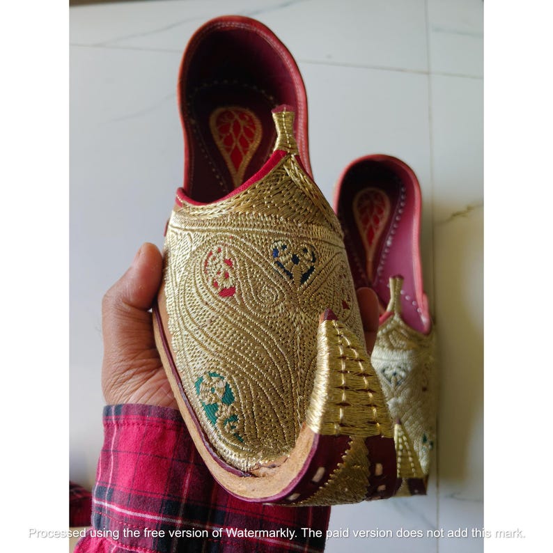 May include: A pair of gold-colored slip-on shoes featuring intricate metallic embroidery in paisley patterns with small red, blue, and green accents. The shoes have a pointed, upturned toe, a deep red interior lining, and a structured, flat sole design.