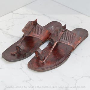 May include: A pair of dark brown leather sandals featuring a wide strap with horizontal embossed lines, a central braided cord, and a toe loop. The flat soles have stitched edges and a pointed heel section, showcasing a weathered, textured finish.