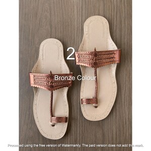 Op de afbeelding: Een paar platte sandalen met een lichtbeige zool met decoratieve stiksels en een metallic bronskleurige bovenband. Het ontwerp bevat een gevlochten teenstuk en een brede band met ingewikkelde uitgesneden patronen. Tekst leest 2 Bronze Colour.