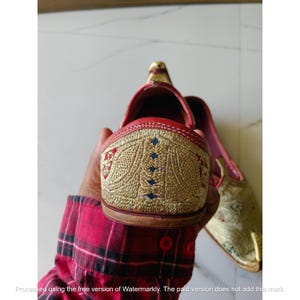 May include: A pair of slip-on shoes featuring a pointed toe and a red leather trim. The exterior is decorated with intricate gold-colored embroidery in a floral pattern, with small blue accents integrated into the stitching on the heel counter.
