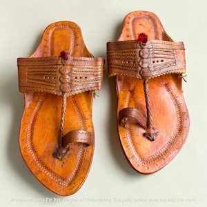 May include: A pair of tan leather sandals featuring a wide strap with intricate embossed patterns, a central braided post, and a small red thread accent at the top. The footwear has a rounded toe design with visible stitching along the edges.
