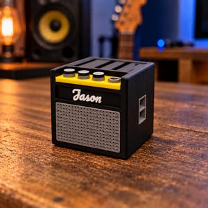 Puede incluir: Un altavoz negro en forma de cubo diseñado para parecerse a un amplificador de guitarra. Cuenta con un panel superior amarillo con tres perillas de control circulares, una rejilla frontal de malla gris y el nombre Jason escrito en blanco.