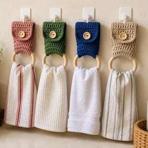 Op de afbeelding: Vier hangende keukendoeken, elk met een gehaakte bovenkant in oudroze, bosgroen, marineblauw of beige, vastgezet met een houten knoop. Elke handdoek hangt aan een gladde houten ring bevestigd aan een witte muurhaak.