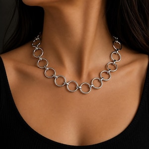 Puede incluir: Un collar de cadena en tono plateado compuesto por una serie de anillos metálicos circulares uniformes y entrelazados. El collar presenta un acabado pulido y reflectante, creando un patrón continuo de eslabones conectados alrededor del cuello.