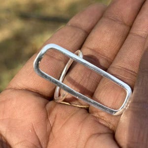 Puede incluir: Un anillo de plata con un marco rectangular grande y alargado que se extiende verticalmente sobre el dedo. La banda es delgada y circular, conectándose a la base del rectángulo de metal liso y pulido que se asienta sobre el dedo.