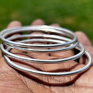 Op de afbeelding: Een set van drie dunne, cirkelvormige zilverkleurige armbanden met een gladde, gepolijste afwerking. De armbanden zijn op elkaar gestapeld en hebben een eenvoudig, afgerond profiel en een reflecterend metalen oppervlak dat het licht vangt.