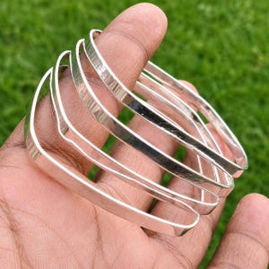 Könnte beinhalten: Ein Set aus vier silberfarbenen Armreifen mit quadratischem Design. Jeder Armreif verfügt über eine glatte, reflektierende Metalloberfläche und eine gleichmäßige Breite, wodurch ein stapelbares Schmuckset mit klaren, geometrischen Linien entsteht.