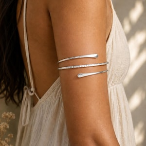 May include: A silver-toned arm cuff featuring a triple-wrap spiral design. The piece consists of two smooth, tapered ends and a central band with a textured, hammered finish, creating a continuous coiled appearance around the upper arm.