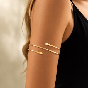 May include: A gold-toned metal arm cuff featuring a double-band design that wraps around the upper arm. The piece has a smooth, polished finish with a slightly textured section on one of the bands and rounded, tapered ends for a clean look.