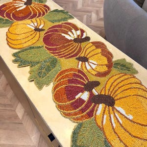 Puede incluir: Un camino de mesa rectangular con un diseño de cuentas que muestra varias calabazas en tonos naranja, amarillo y rojo oscuro, acentuado con hojas de cuentas verdes. Las calabazas tienen tallos marrones y reflejos blancos texturizados.
