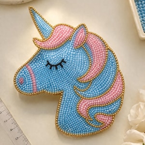 May include: A beaded unicorn patch featuring a light blue face and pink mane, outlined in gold beads. The unicorn has a closed eye with black lashes and a horn. A clear plastic ruler is visible, showing the patch is approximately 3.5 inches tall.
