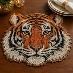 May include: A tiger-shaped placemat crafted from intricate beadwork in shades of orange, black, white, and gold. The design features a detailed tiger face with piercing eyes, a pink nose, and a scalloped edge that follows the animal's silhouette.