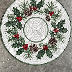 May include: A round beaded placemat featuring a white background with a circular wreath design. The wreath consists of green holly leaves, bright red berries, and brown pinecones. The outer edge is finished with a thin green beaded border.