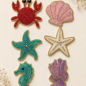 Op de afbeelding: Een set van zes geborduurde patches met zeedieren en schelpen. De set bevat een rode krab, een roze schelp, een groenblauwe zeester, een witte zeester, een groenblauw zeepaardje en een paars zeepaardje, allemaal omlijnd met gouden kralen.