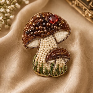 May include: A handcrafted mushroom brooch featuring intricate beadwork. The cap is adorned with brown beads and a single faceted red crystal. The stem is covered in cream-coloured seed beads, while the base includes green glass bugle beads.